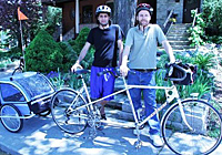 Two men hold up a tandem bike with a small trailer, ready for a bike ride for charity for Rifton tricycles