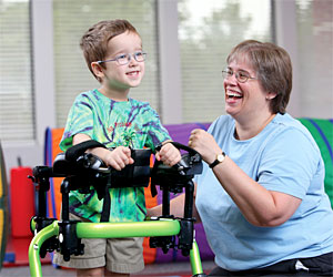 A young boy movement disorders is led by a smiling therapist as he walks in a Rifton gait trainer.