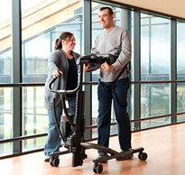 A smiling therapist at a rehab facility helps an adult patient with complex disabilities walk in a TRAM device