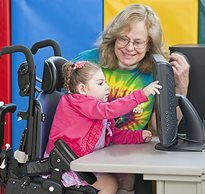 A teacher smiled at a special needs student seated in a adaptive chair practicing motor skills on a computer screen