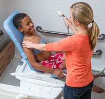 A boy with special needs smiles in his Rifton Wave bath chair while a caregiver assists him