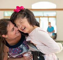 A caregiver and young girl in a Rifton Mobile Stander laugh together while posing for a portrait.