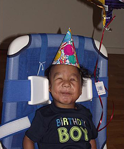 A young boy living with Trisomy is supported by his Rifton Bath Chair with balloons attached, while he celebrates his first birthday in a party hat