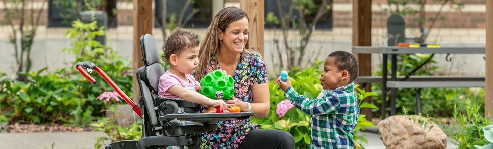 A physical therapist talks to a little boy and a little girl standing in a Rifton Stander.
