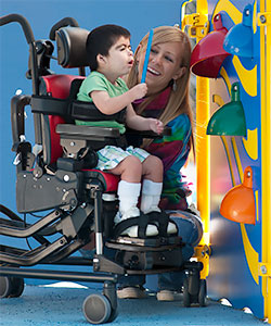 A child using adaptive seating reaches to tap on a colorful bell, while a young woman looks on smiling