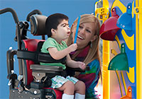 A child using adaptive seating reaches to tap on a colorful bell, while a young woman looks on smiling