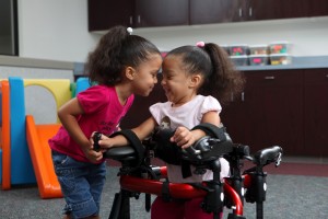 A smiling Ella in her red gait trainer recieves encouragement from her sister as she works on early mobility
