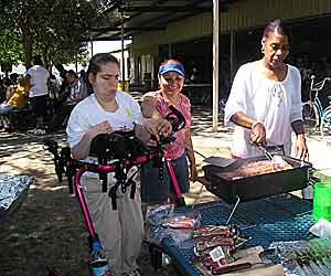 An adult standing in a gait trainer shows the benefits of movement disorder treatment – she helps cook food on an oudoor grill