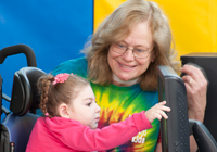A Young Girl in an Activity Chair Pushes a Screen with the Aide of a RTS Device While a Smiling Woman Assists Her