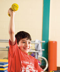 A young boy with cerebral palsy exercises by lifting a yellow hand weight up in the air