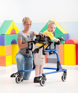 A woman therapist helps a young boy with disabilities into a Pacer gait trainer