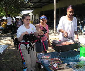 A disabled woman supported by a gait trainer, flips burgers on the bbq, helped by her Life Services personal advocate