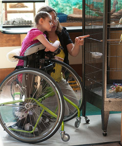 A woman points to a bird in a cage while a young girl in a stander looks on
