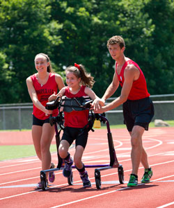 A young girl with cerebral palsy is guided by her two teams in a gait trainer as she runs the straightaway of an outdoor track
