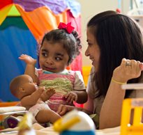 A therapist and daughter in a playroom, both smiling, as the little girl waves while playing with a doll