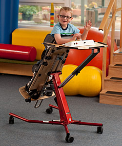 A young boy in a classroom on an supported standing device plays with a toy 