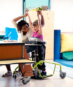 A classroom setting with a therapist and a young girl in a Dynamic Stander raising their hands up cheering and smiling showing the benefits of standing.