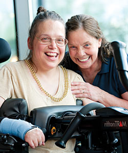 Joyce Checksfield leans over Kandi Elmore’s shoulder as she sits in a Rifton Tram and they both smile for the camera