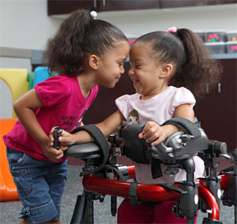 A little girl in an assistive technology device smiles at her sister who leans in close to her face.
