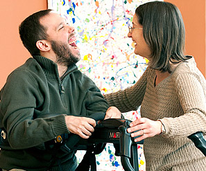 A man laughs and smiles with delight as a caregiver helps him transfer to a standing position