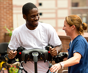 A caregiver aids a young patient in a gait trainer in a courtyard as they smile at one another