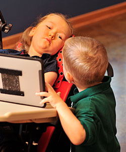 A young boy interacts with his sister seated in an activity chair by using a supplemental aid and service tablet device