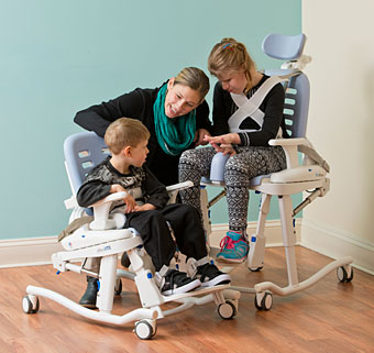A young girl and boy sit in the new Rifton Hygiene and Toileting System (HTS) while their caregiver chats with them.