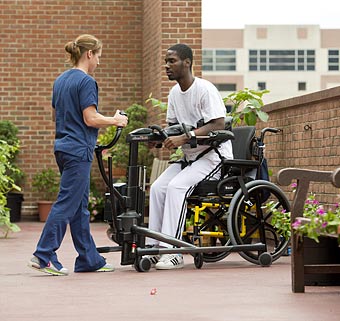 A member of the ASPHP assists a patient from a wheelchair to standing position using the Rifton TRAM outside in the garden of a hospital.