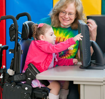 A teacher smiles at a child with cerebral palsy who is touching a computer screen from her adaptive chair