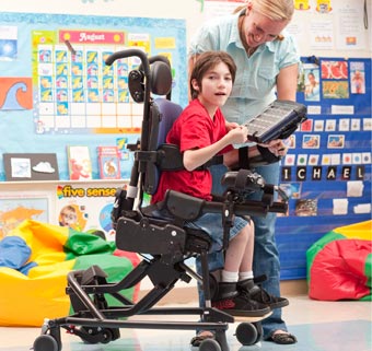 In a classroom setting, a young boy with cerebral palsy is seated in an adaptive chair while his teacher holds up a game for him to play with 