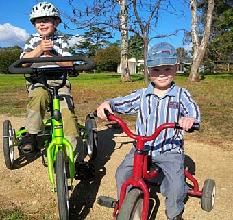 Two brothers smiling at the camera are outside on adaptive tricycles with blues skies in the background
