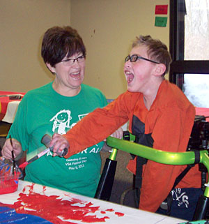 A student with special needs using an adaptive gait trainer smiles big as he paints red and blue on a piece of paper with the assistance of his teacher.