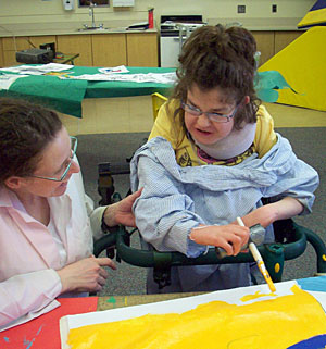 A girl with special needs uses adaptive painting equipment and a gait trainer to paint yellow onto a sheet of paper with the assistance of her teacher.