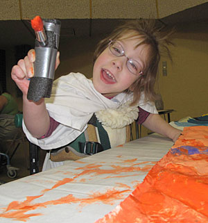 A child with disabilities in a Dynamic stander holds out her adaptive paint brush and smiles at the camera.