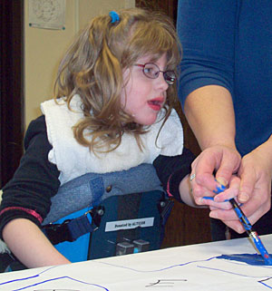 A young child with disabilities in an adaptive stander gets assistance from her teacher to paint a figure in blue on the paper.