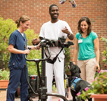 Two therapists using SPHM technique aid a patient in a Rifton TRAM as they smile at the guide dog assisting them all
