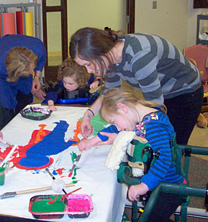 A group of students with special needs using adaptive equipment around a table paint colorful figures with the help fo their therapist and teacher.