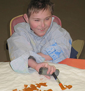 A boy with special needs smiles sitting in an activity chair as he holds an adaptive paint brush coloring yellow on the paper.