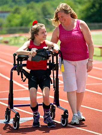 A therapist assists a young girl walking in a gait trainer as she works on motor skills, both smiling at the positive outcomes of the training session at the track