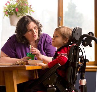 At a kitchen table, a mother with her special needs child in an adaptive seating device 