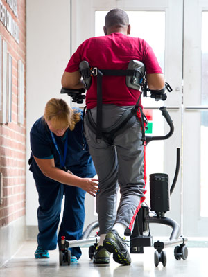 A neurological rehab specialist assists a patient practicing early mobility in a TRAM device by walking down the hallway
