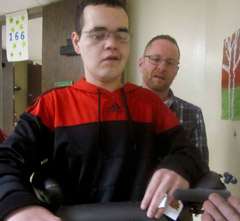 A TBI patient positioned properly using the chest support in the TRAM walks down the hallway as his teacher looks on.