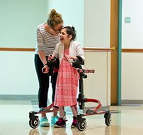 A young girl smiles as she uses a Rifton Pacer gait trainer with guidance from her therapist
