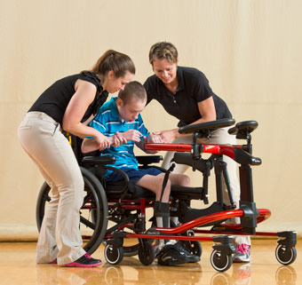 Two therapists help a young boy transition from a wheelchair to the dynamic gait trainer, new adaptive equipment that will help him improve mobility
