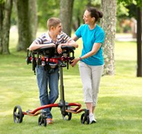 A therapist helps guide a young boy in a Rifton Pacer gait trainer as he walks on the grass in a park.