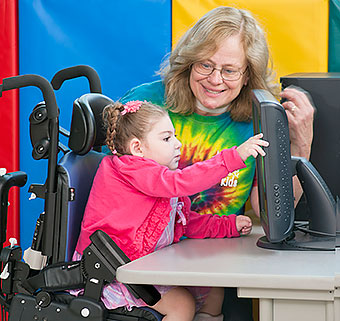 A teacher smiled at a special needs student seated in a adaptive chair practicing motor skills on a computer screen