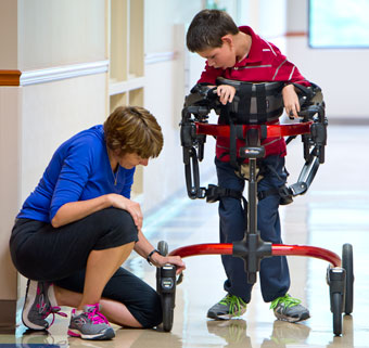 A therapist stops to change a setting on a gait trainer as she works with a child in the school hallway