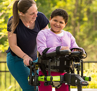 A therapist with her arms around a child with disabilities smiles as she practices dynamic movement in her gait trainer