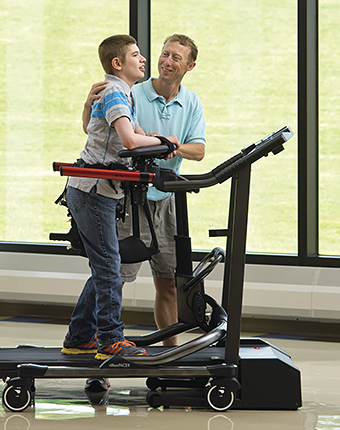 A teacher helps a student in the classroom with ambulation using a treadmill set-up with a gait trainer