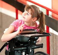 A young girl positioned in a standing mobility device points to her communication tray.